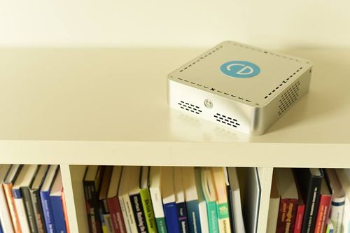 A photograph of a 20-centimetre cuboid aluminium home server box. It rests in a minimalist fashion on top of a white bookshelf with no wires.